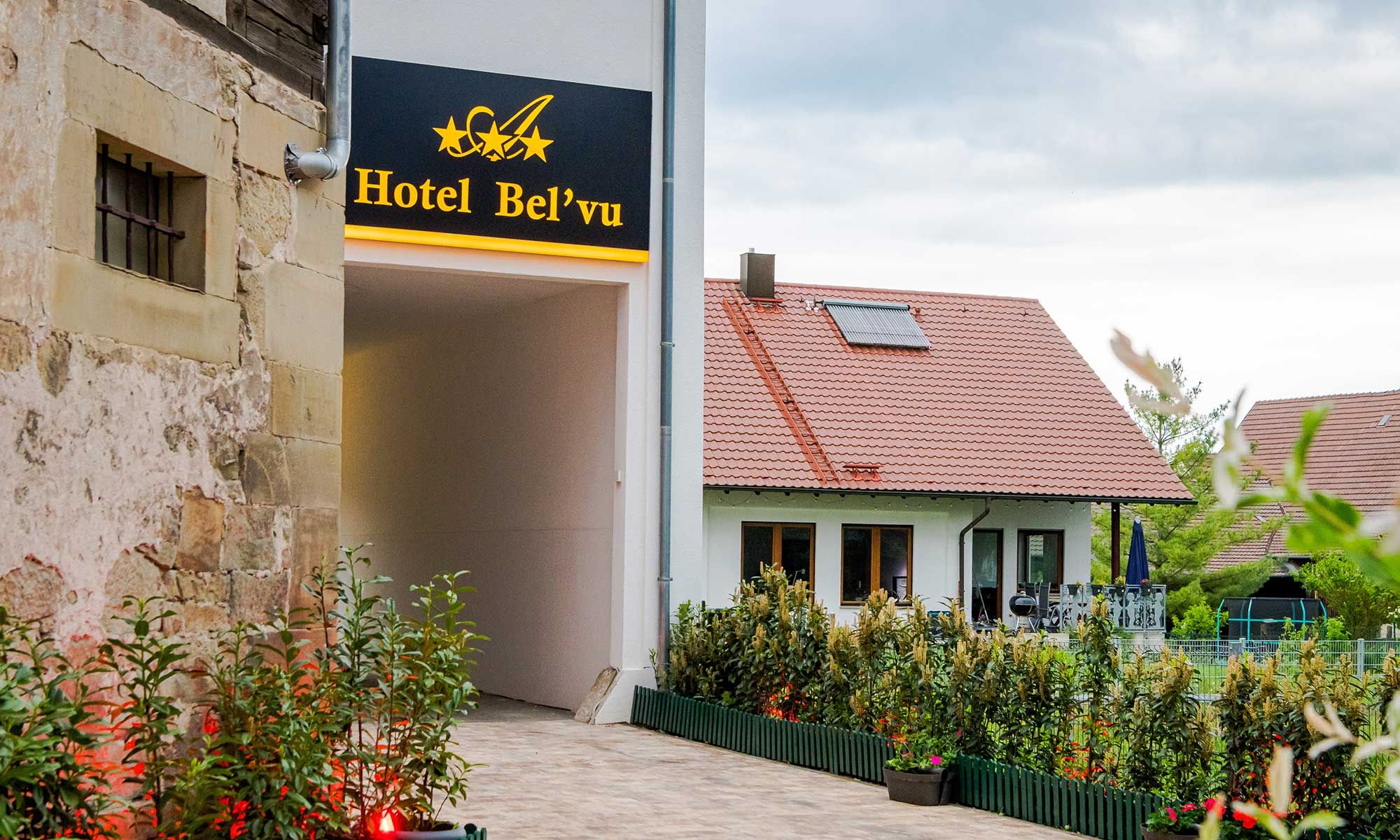 Entrada de hotel com uma placa indicando 'Hotel Bel'vu' e um edifício tradicional com telhado de telhas vermelhas ao fundo.