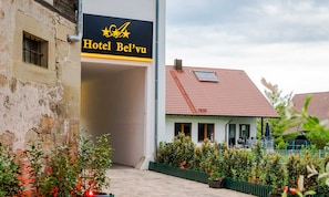 Entrada de un hotel con un letrero que dice 'Hotel Bel'vu' y un edificio tradicional con techo de tejas rojas al fondo.