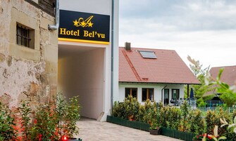 Entrada de un hotel con un letrero que dice 'Hotel Bel'vu' y un edificio tradicional con techo de tejas rojas al fondo.