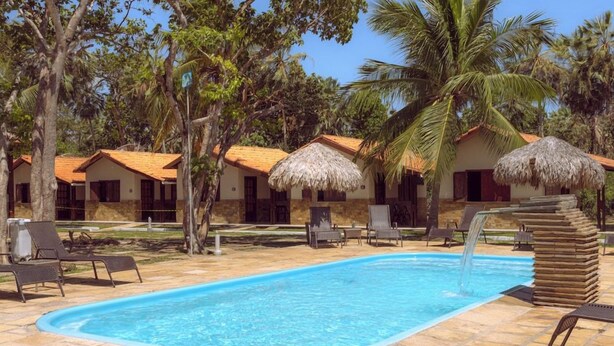 Piscina com espreguiçadeiras e guarda-sóis de palha, cercada por palmeiras e edifícios com telhados de azulejos.
