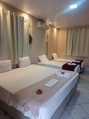 A hotel room with three beds, each with white linens and a red flower decoration. There is an air conditioning unit mounted on the wall, and the room has a brick wall design. - Orla Náutica (Barreirinhas)
