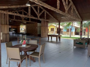 An outdoor covered area with a ping pong table, chairs, and a dining table with a vase of flowers. - Orla Náutica (Barreirinhas)