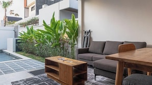 A modern living room with a grey sofa, a wooden coffee table, and a dining table. There is a pool in the background and a balcony with plants. - Cypress Villa by Zuzu (Ungasan)