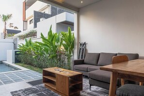 Een moderne woonkamer met een grijze bank, een houten salontafel en een eettafel. Achtergrond is een zwembad en er is een balkon met planten.