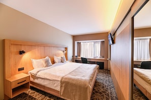 Quarto de hotel com cama, mesa de cabeceira, escrivaninha, cadeira e televisão fixada na parede.