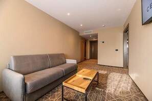 Un moderno vestíbulo de hotel con un sofá gris, una mesa de centro de madera, alfombra estampada y una puerta que da a un pasillo.
