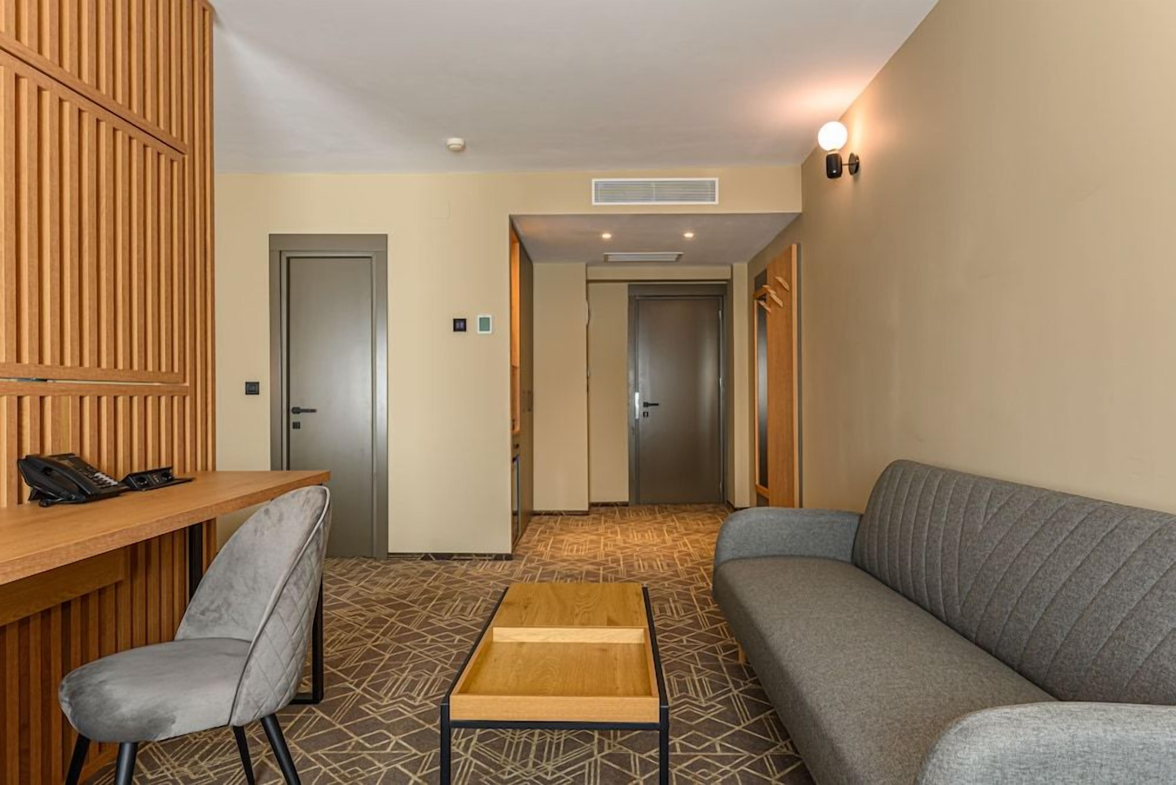 A hotel room with a wooden reception desk, a grey sofa, a small wooden table, and a patterned carpet.