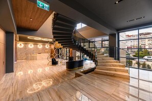 Um hall de hotel moderno com piso de mármore, balcão de recepção, escadaria e amplas janelas com vista para a cidade.