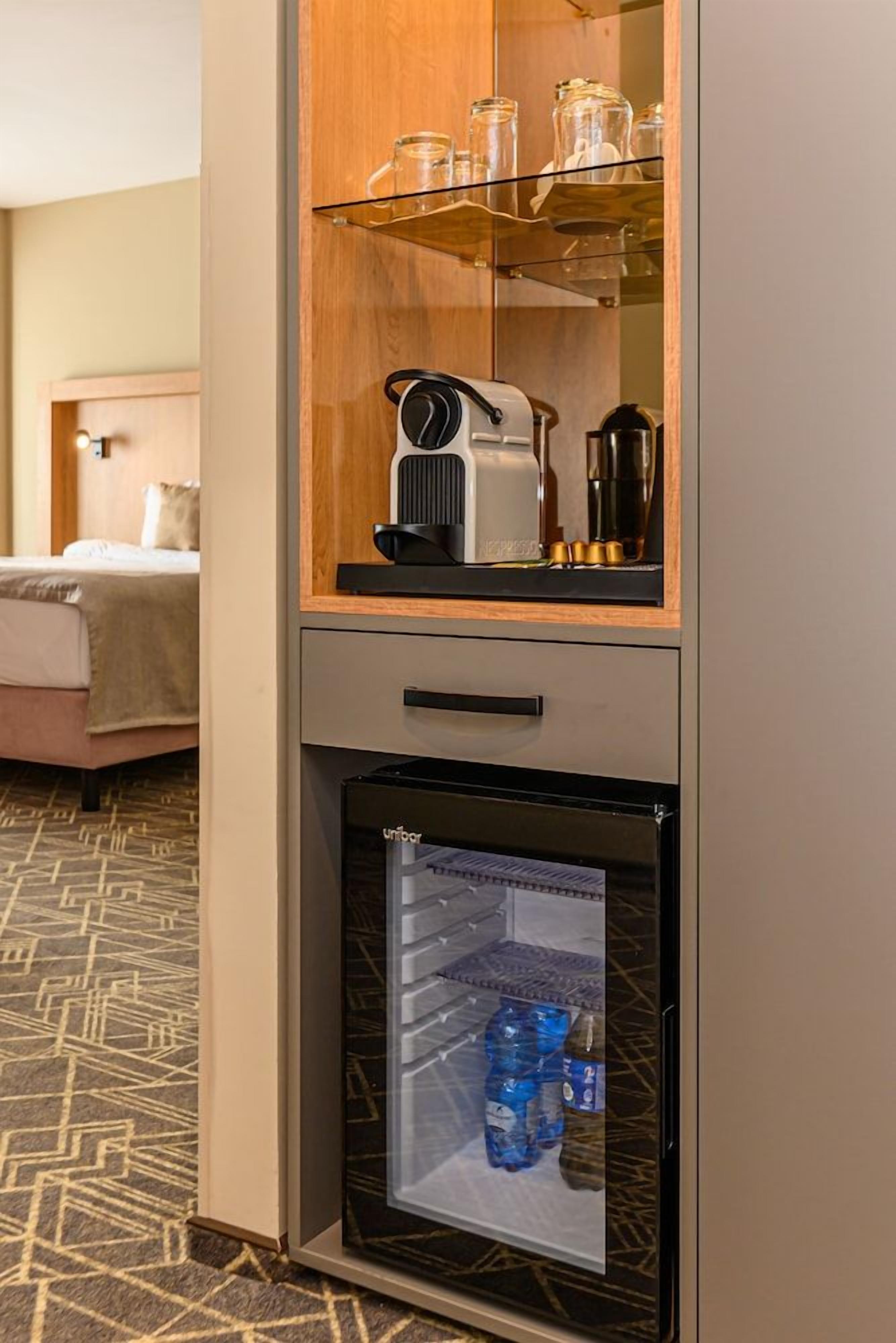 A minibar with a coffee maker, a glass shelf with jars, and a refrigerator with bottled water.