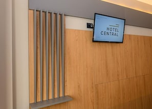 Zona de recepción de hotel con pared de paneles de madera, un televisor montado mostrando 'HOTEL CENTRAL' y un reloj digital que indica la hora.