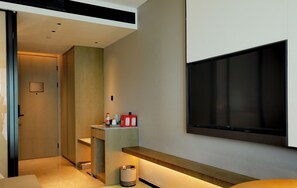A modern hotel room with a flat-screen TV mounted on the wall, a small table with a trash bin, and a wooden bench.