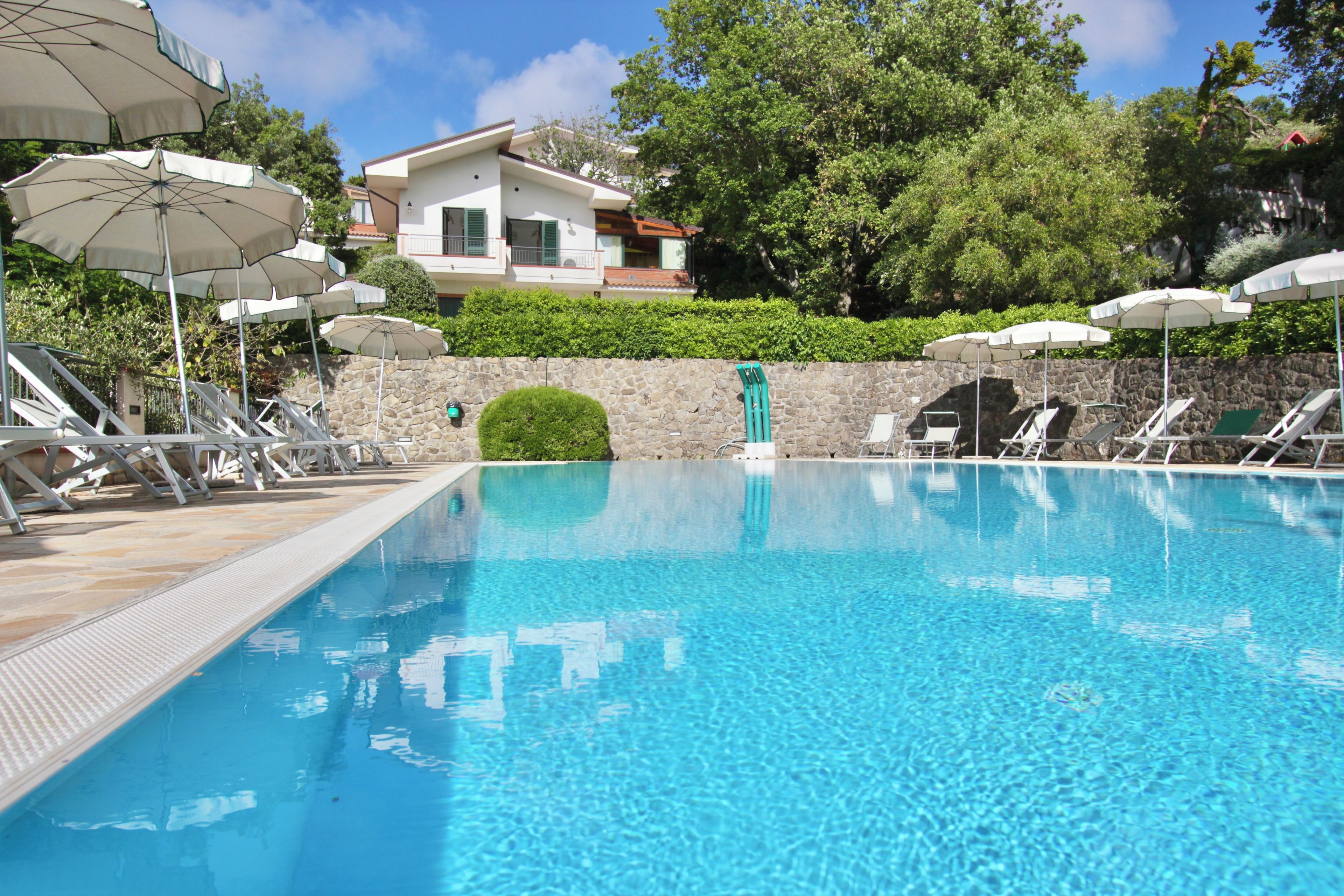 A clear swimming pool with lounge chairs and umbrellas, surrounded by a stone wall and greenery.