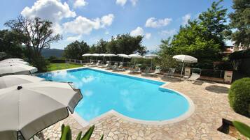 A swimming pool with lounge chairs and umbrellas, surrounded by greenery and a clear sky.