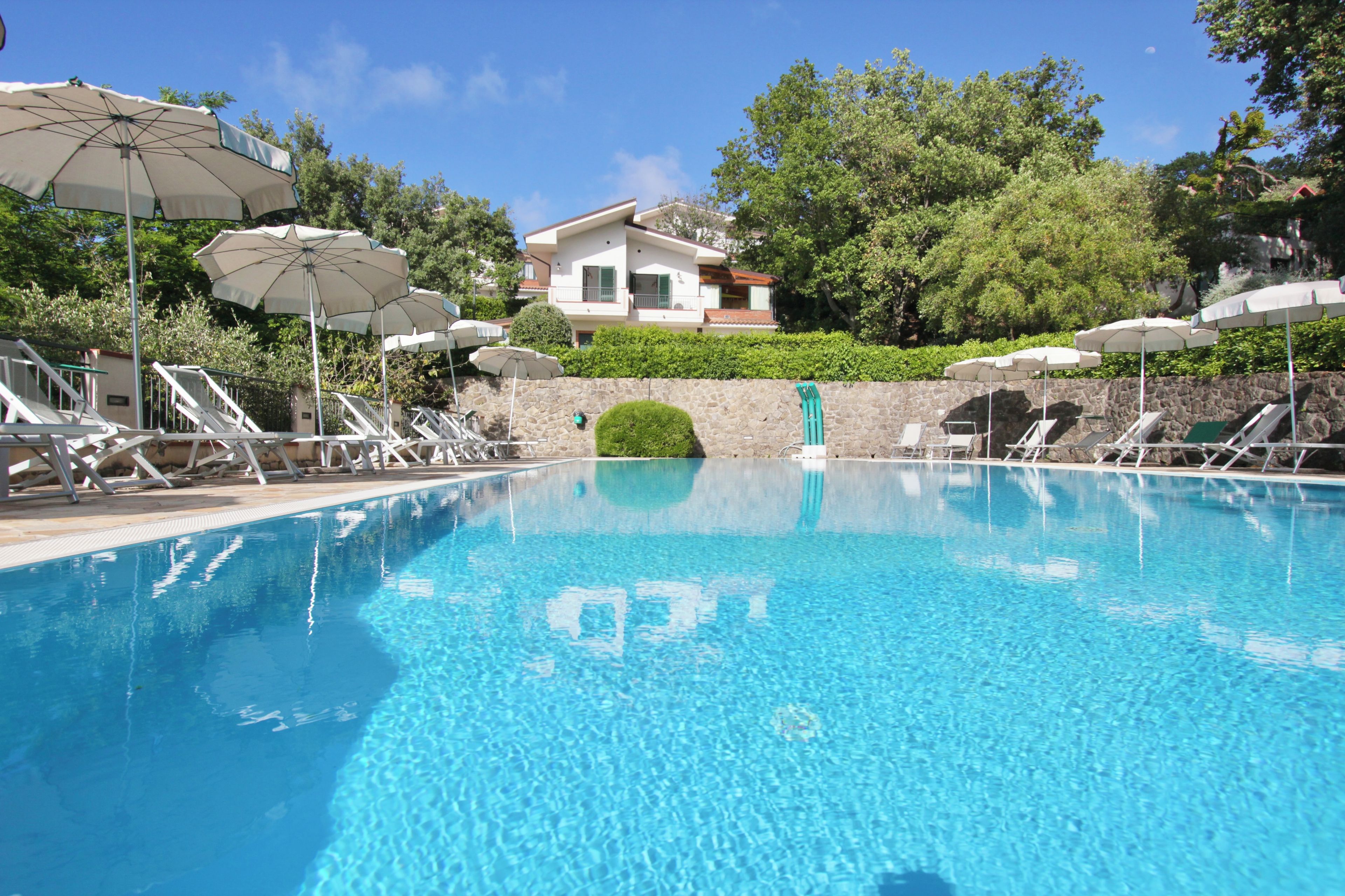 Une piscine entourée de chaises longues et de parasols, avec de la verdure tout autour et un bâtiment en arrière-plan.