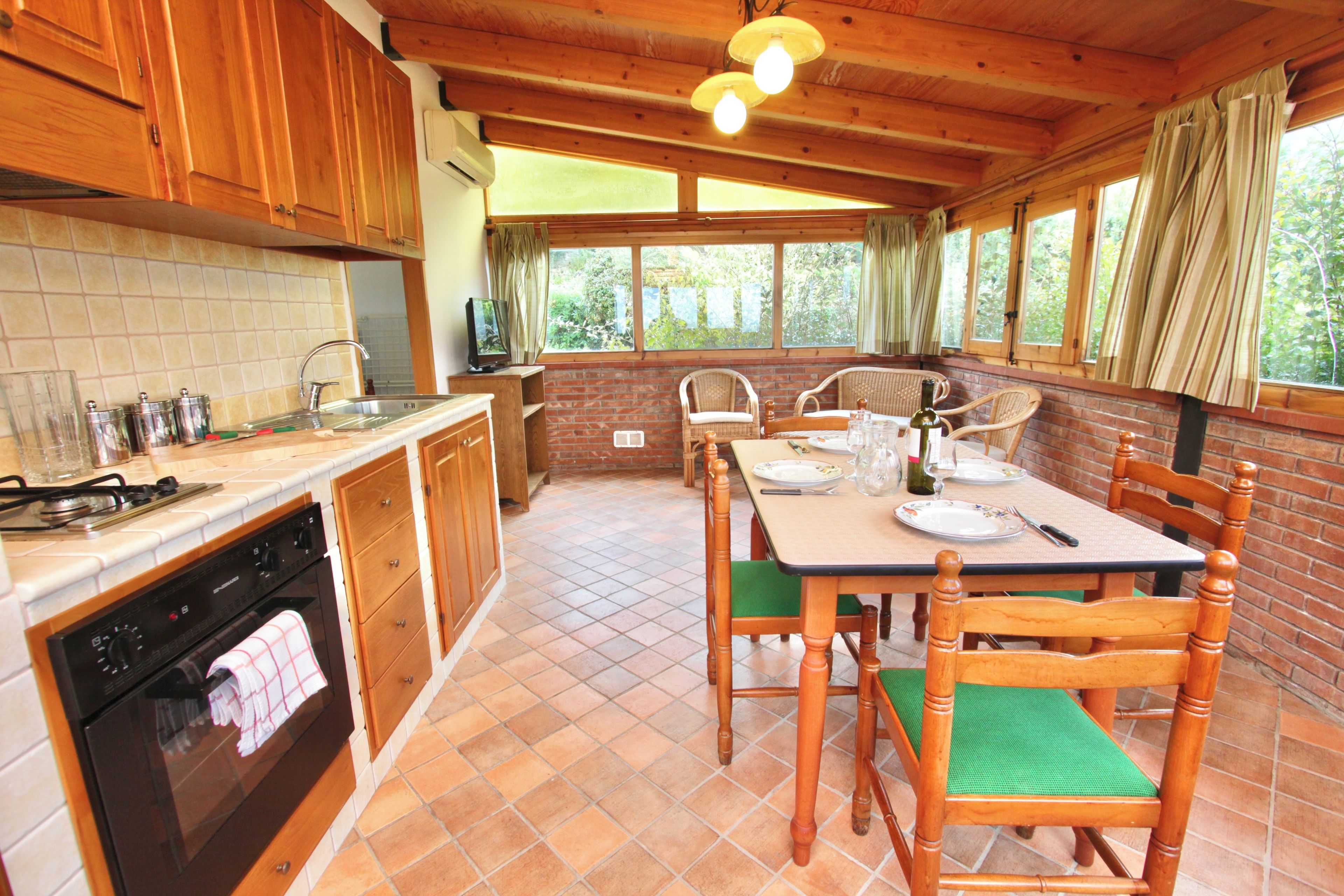 Une cuisine avec des meubles en bois, une table à manger avec des chaises et une vue sur la verdure par les fenêtres.