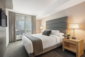 Une chambre d’hôtel avec un grand lit, une table de chevet avec une lampe, un téléviseur fixé au mur et une fenêtre avec des rideaux.