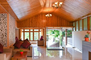 A spacious room with wooden ceiling and walls, a seating area with cushions, a glass door leading outside, and a chandelier.