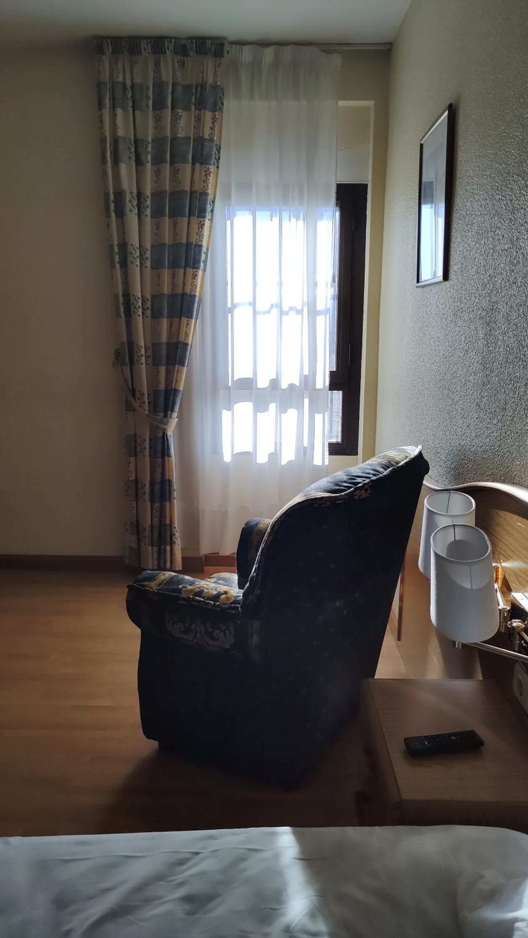 Habitación de hotel con una cama individual, una silla, una mesita de noche con lámpara y una ventana con cortinas.