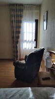 Habitación de hotel con una cama individual, una silla, una mesita de noche con lámpara y una ventana con cortinas.