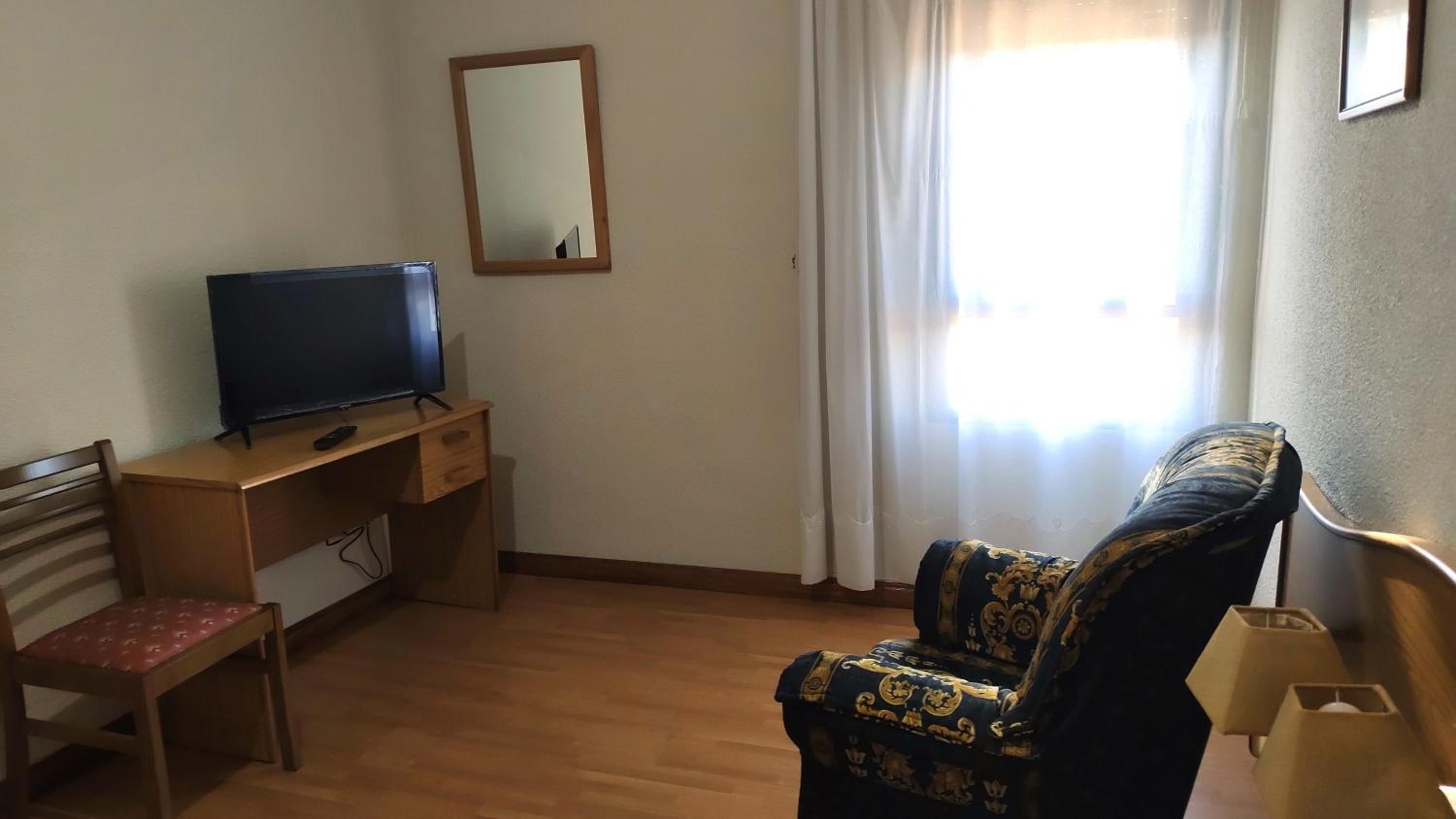 Una habitación con un escritorio de madera, una televisión, un sillón estampado y una ventana con cortinas.