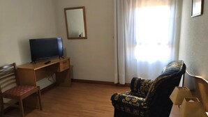 Ein Zimmer mit einem Holztisch, einem Fernseher, einem gemusterten Sessel und einem Fenster mit Vorhängen.