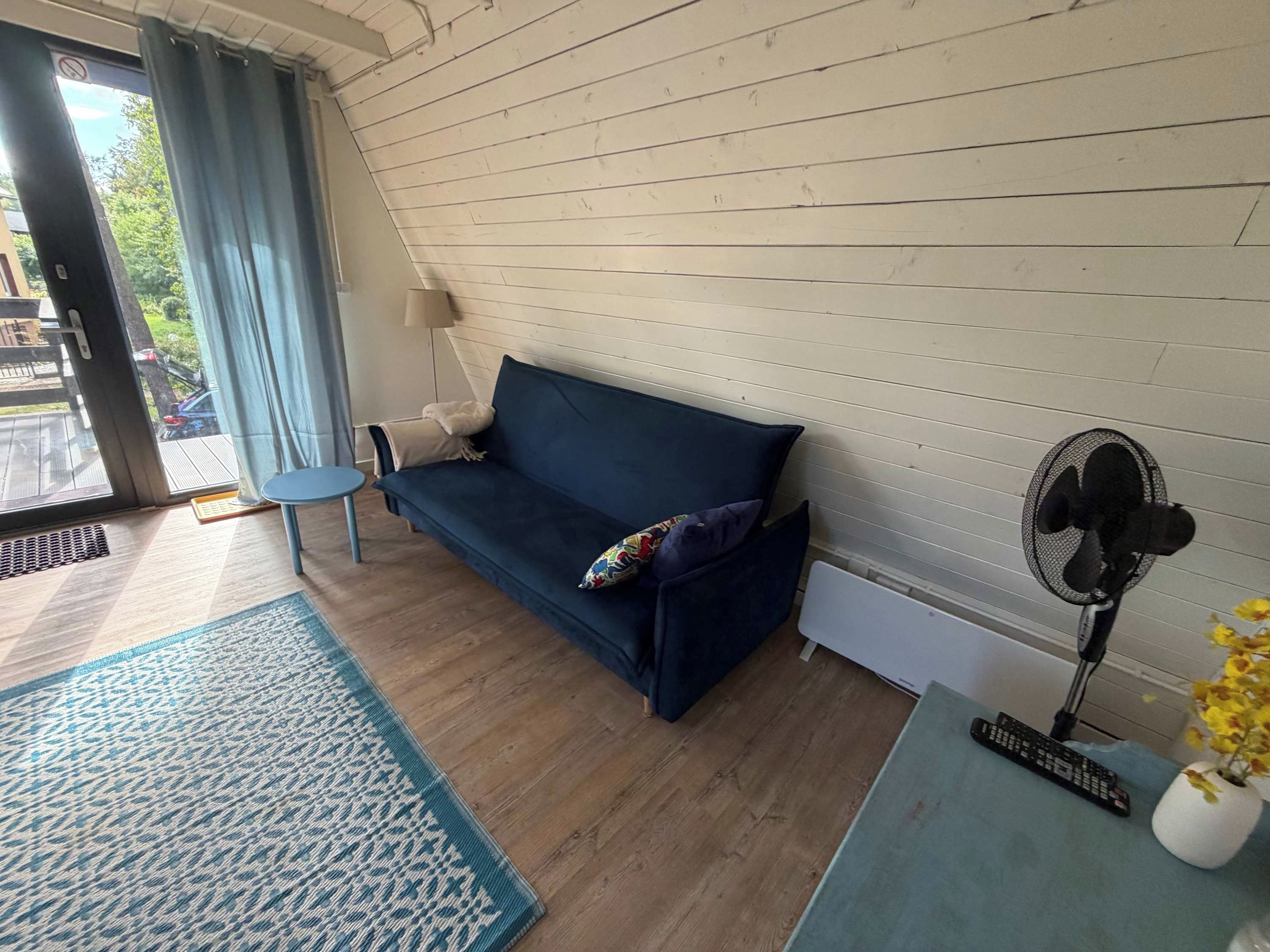Una habitación con un sofá azul, una mesita, un ventilador de pie y un control remoto sobre una mesa.