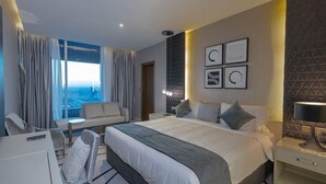 A hotel room with a large bed, a sitting area with a chair, a desk, and a view of the city through large windows. - Hotel Suites - Damac Tower Riyadh (Riyadh)