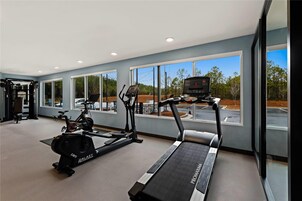 Un gimnasio con cinta de correr, elíptica y máquinas de pesas, ventanas amplias y vista al estacionamiento.