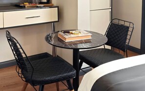 Ein kleines Zimmer mit einem Bett, einem runden Tisch mit zwei Stühlen, einem Buch und einer Lupe auf dem Tisch.