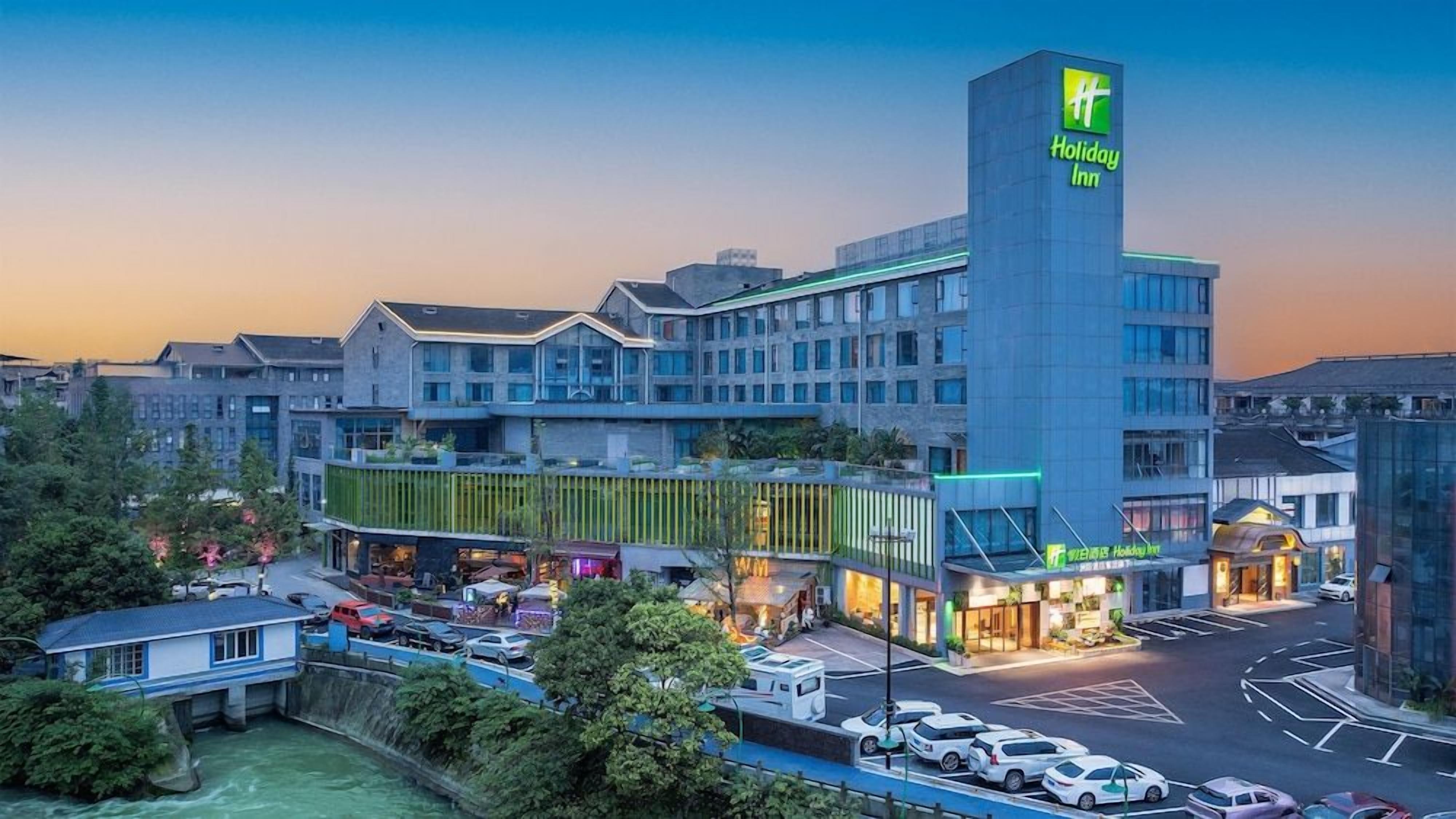 A Holiday Inn hotel with a modern design, featuring a prominent vertical tower with the logo, a spacious parking area, and a well-lit entrance.