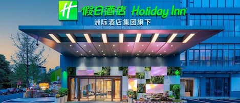 A Holiday Inn hotel entrance with a modern design, featuring a prominent logo, a covered entrance area, and a landscaped front garden.