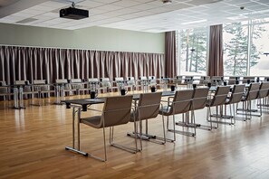 Sala de conferência com mesas e cadeiras dispostas em fileiras, um projetor e amplas janelas com cortinas.