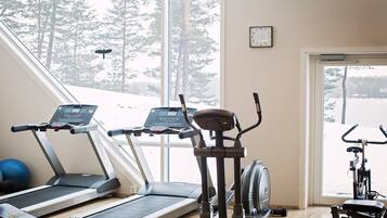 Ett gym med löpband, en crosstrainer och en motionscykel, med utsikt över snötäckta träd utanför.