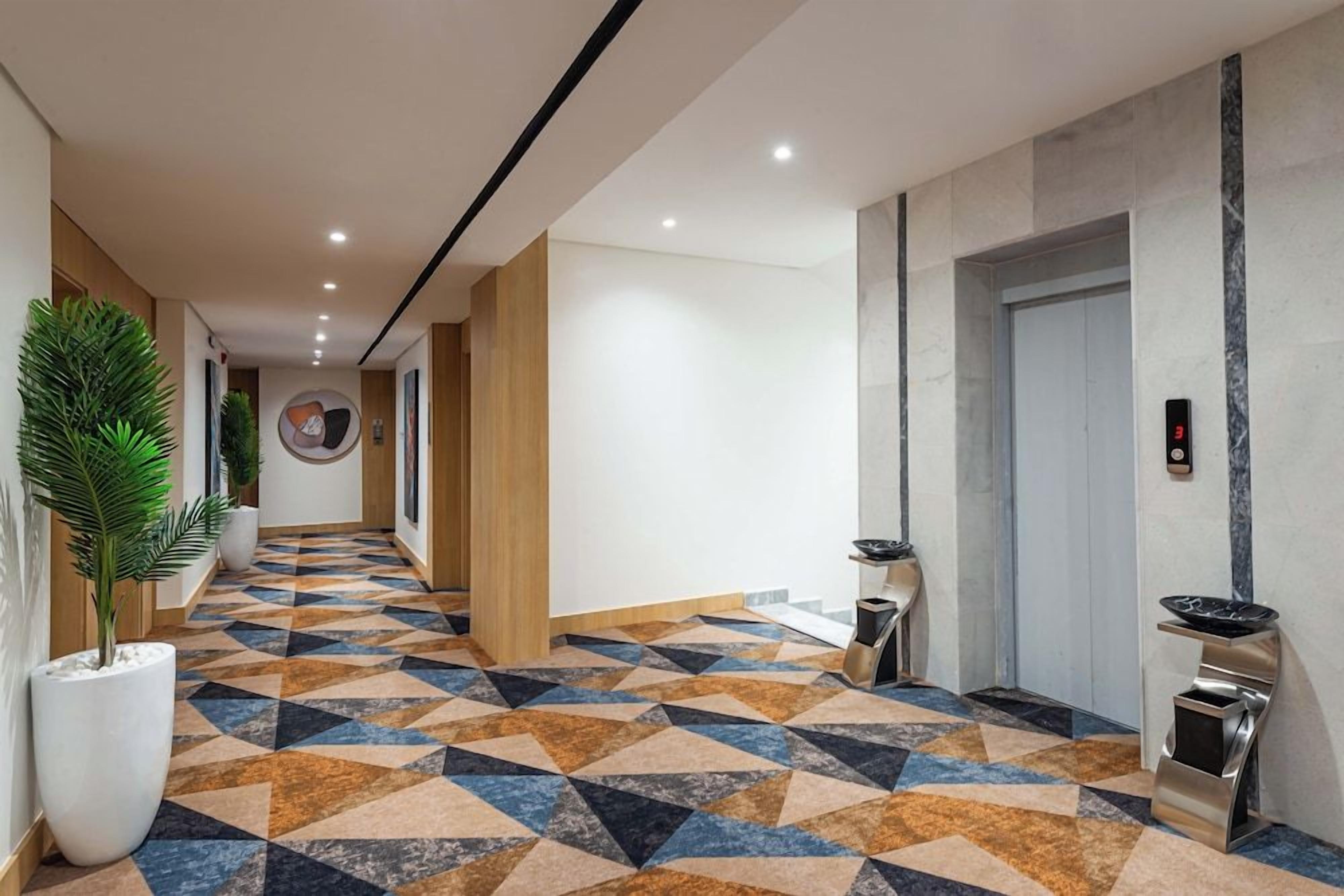 A hotel lobby with a geometric patterned floor, a reception desk, a wall-mounted elevator, and potted plants.