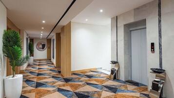 A hotel lobby with a geometric patterned floor, a reception desk, a wall-mounted elevator, and potted plants.
