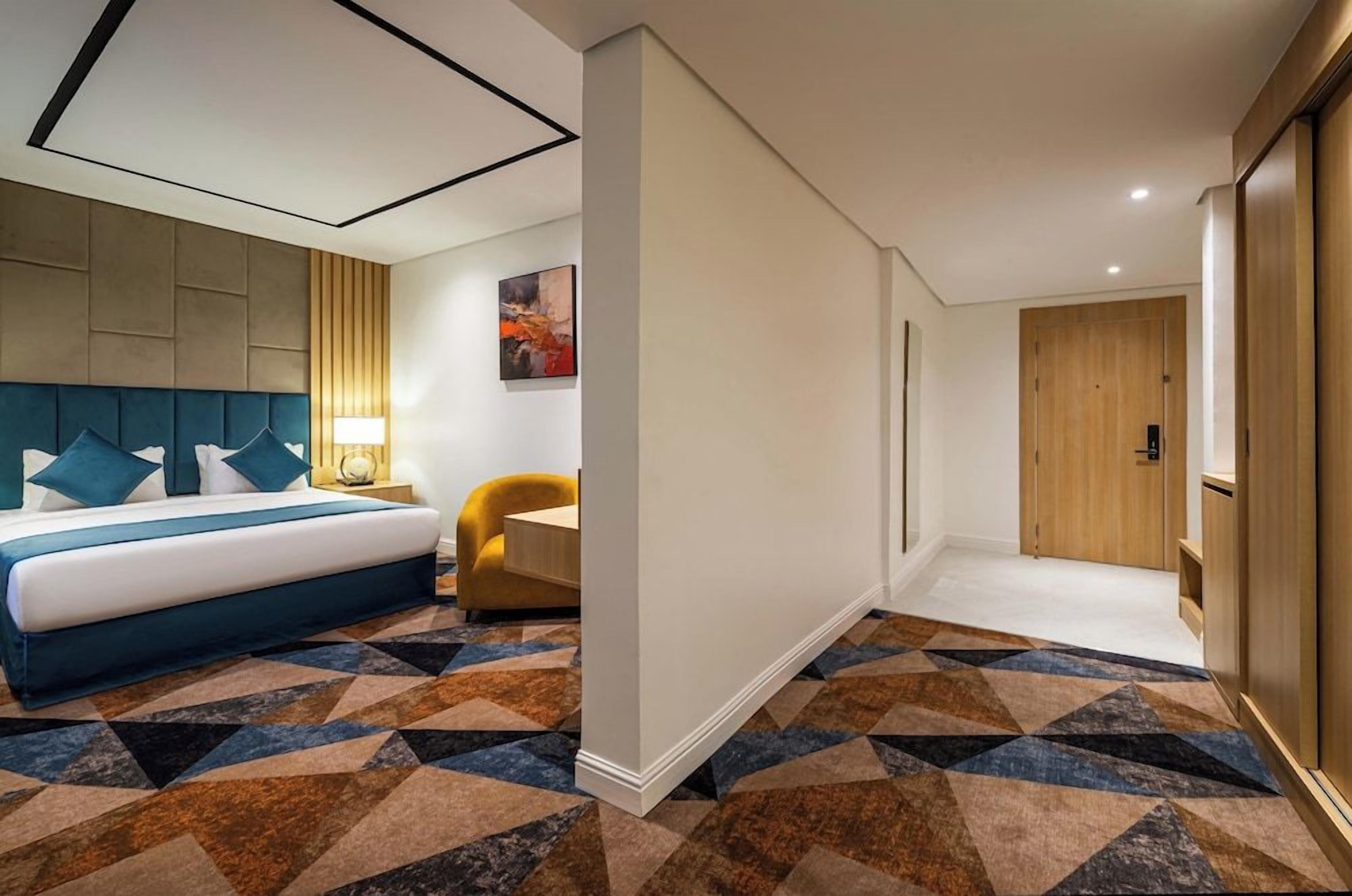 Quarto de hotel com cama, escrivaninha, cadeira, porta e carpete estampado.