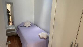Un dormitorio con tragaluz, una cama con colcha morada, un almohadón blanco, un armario y una mesita de noche pequeña.