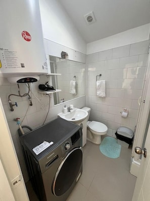 A compact bathroom with a washing machine, sink, toilet, and a mirror. - Departamentos Egaña (Puerto Montt)