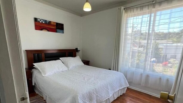 A bedroom with a wooden floor, a bed with white bedding, a hanging light fixture, and a window with sheer curtains. - Departamentos Egaña (Puerto Montt)