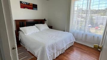 Un dormitorio con piso de madera, una cama con ropa de cama blanca, una lámpara colgante y un ventanal con cortinas.