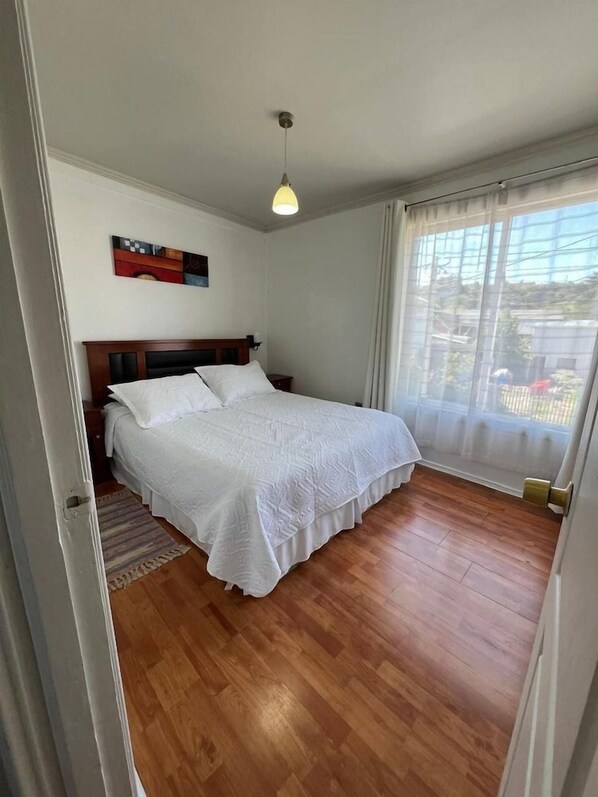 A bedroom with a wooden floor, a bed with white bedding, a hanging light fixture, and a window with sheer curtains. - Departamentos Egaña (Puerto Montt)