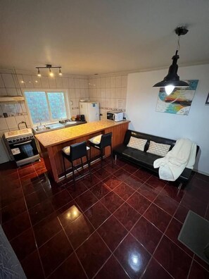 A compact kitchen with a central island, a dining area with a sofa, and a black chair. - Departamentos Egaña (Puerto Montt)