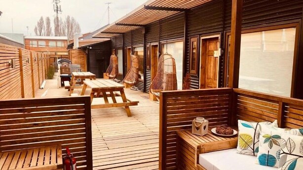 Een houten terras met zitplaatsen en tafels, een bed met kussens, en een raam met uitzicht op een gebouw.