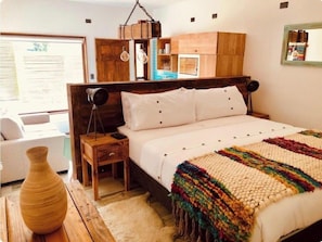 Een slaapkamer met een houten hoofdbord, een witte spreidek en een kleurrijke deken. Er staat een houten nachtkastje met een lamp en een vaas erop.
