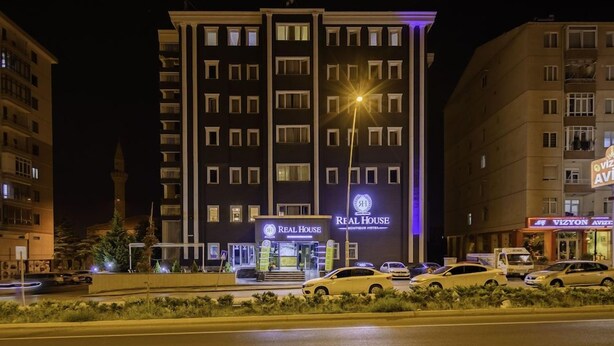 Ein mehrstöckiges Hotel mit auffälligem 'Real House' Schild, einem gut beleuchteten Eingang und Blick auf die Straße mit geparkten Autos.