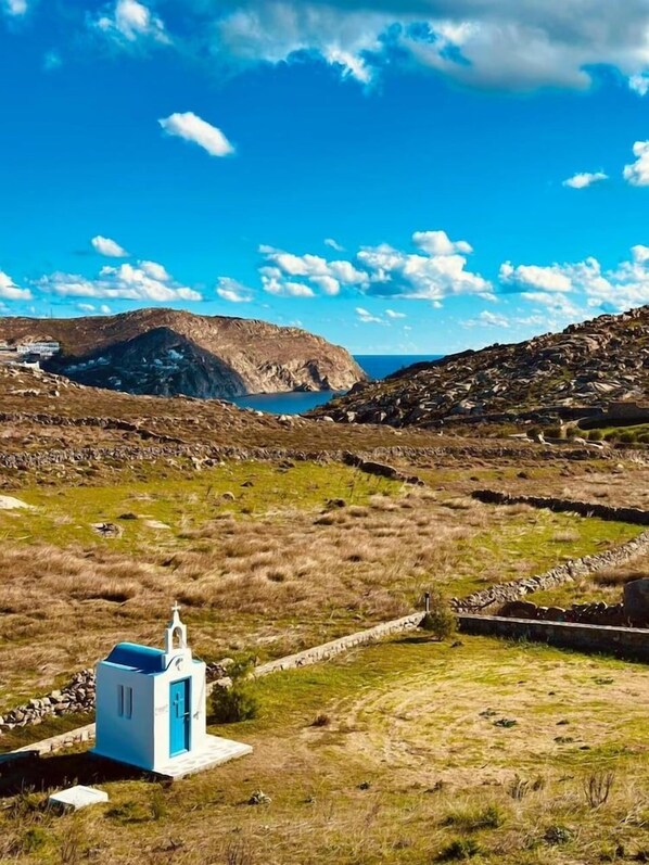 A small white building with a blue door, surrounded by a grassy landscape and rocky terrain. - Anemela Mykonos Villas & Suites (Mykonos)