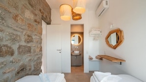A bedroom with two beds, a stone wall, a bathroom with a sink and mirror, and a hanging light fixture. - Anemela Mykonos Villas & Suites (Mykonos)