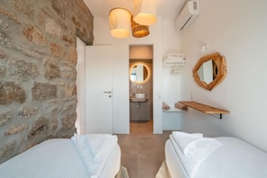 Ein Schlafzimmer mit zwei Betten, einer Steinwand, einem Badezimmer mit Waschbecken und Spiegel sowie einer Pendelleuchte.