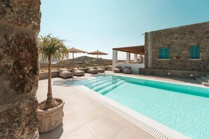 Ein Poolbereich mit Liegestühlen und Sonnenschirmen, eine Steinmauer und Blick auf die Landschaft.