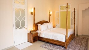Ein Bett mit Baldachin, dessen Holzbettgestell mit weißer Bettwäsche bezogen ist, ein Nachttisch mit Lampe und ein gemustertes Teppich auf dem Boden.